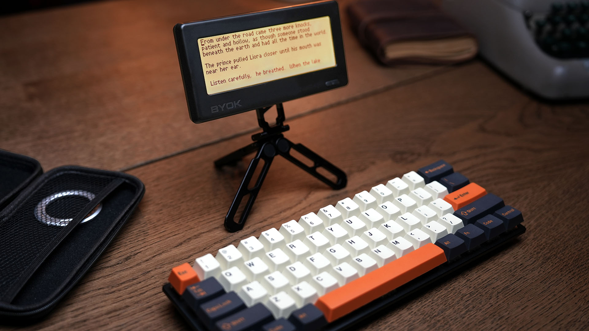 BYOK writing device on its compact ring stand beside a travel case and mechanical keyboard, a draft on the screen